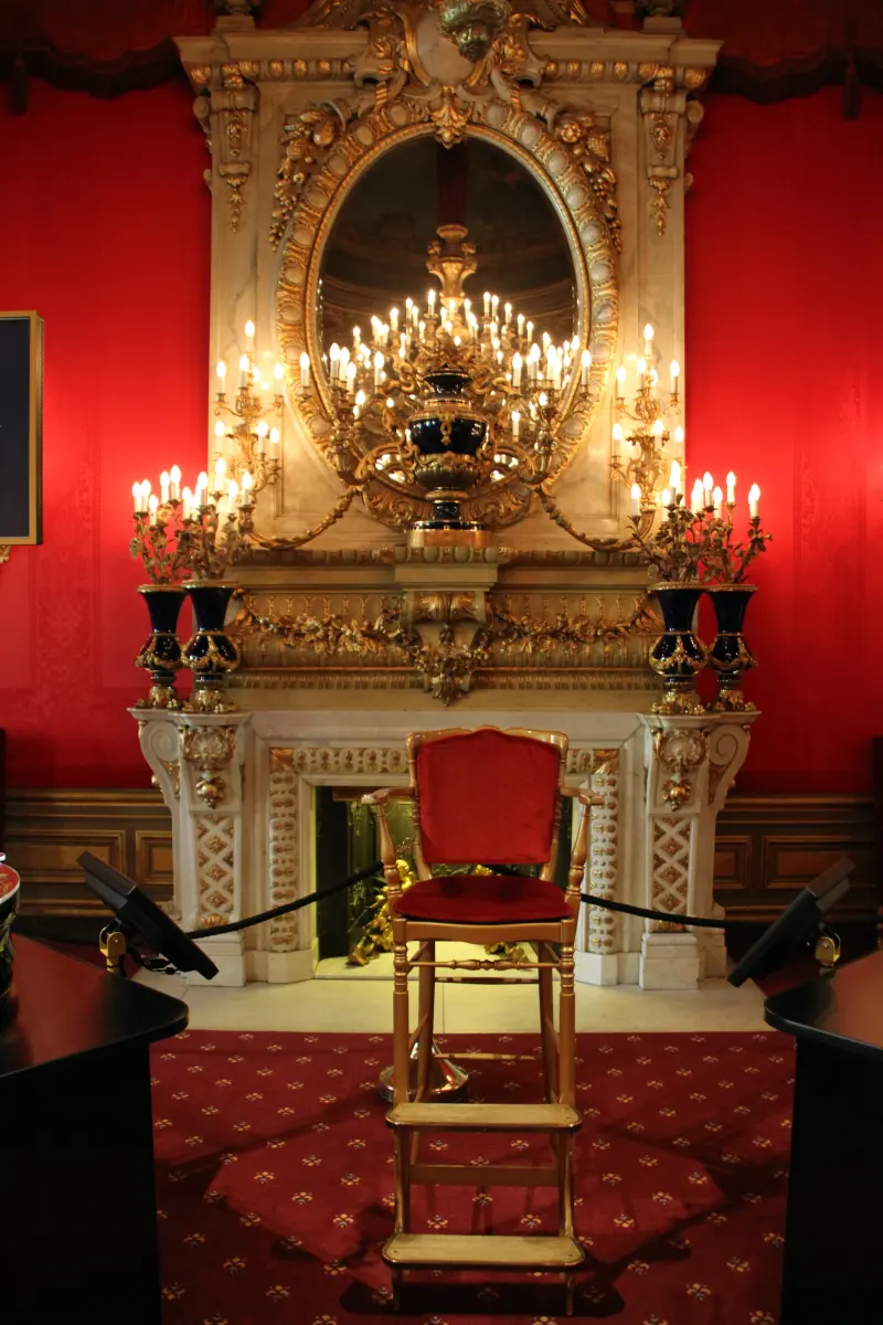 A gaming salon inside the Baden-Baden Kurhaus: scarlet damask walls, a marble chimneypiece under a gilt-framed oval mirror, branched candelabra alight, and the croupier's raised chair on its wooden steps between two gaming tables.