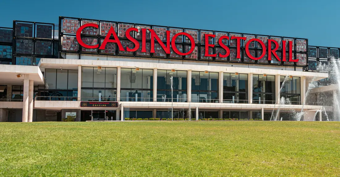 The front of Casino Estoril from the gardens: a long low modernist block, a row of square columns carrying a deep concrete slab over a fully glazed ground floor, with the words CASINO ESTORIL in tall red letters across the roofline and fountains playing to the right.