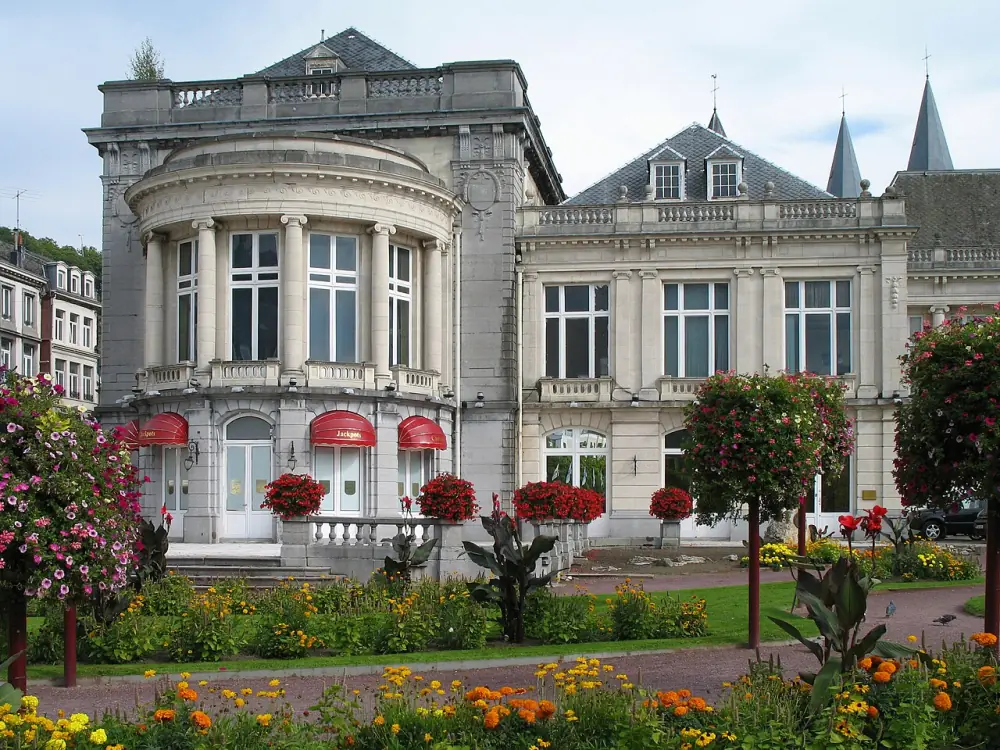 The Casino de Spa seen from the street: a pale stone building with a curved colonnaded bay, mansard roofs and red awnings, flower beds in front.