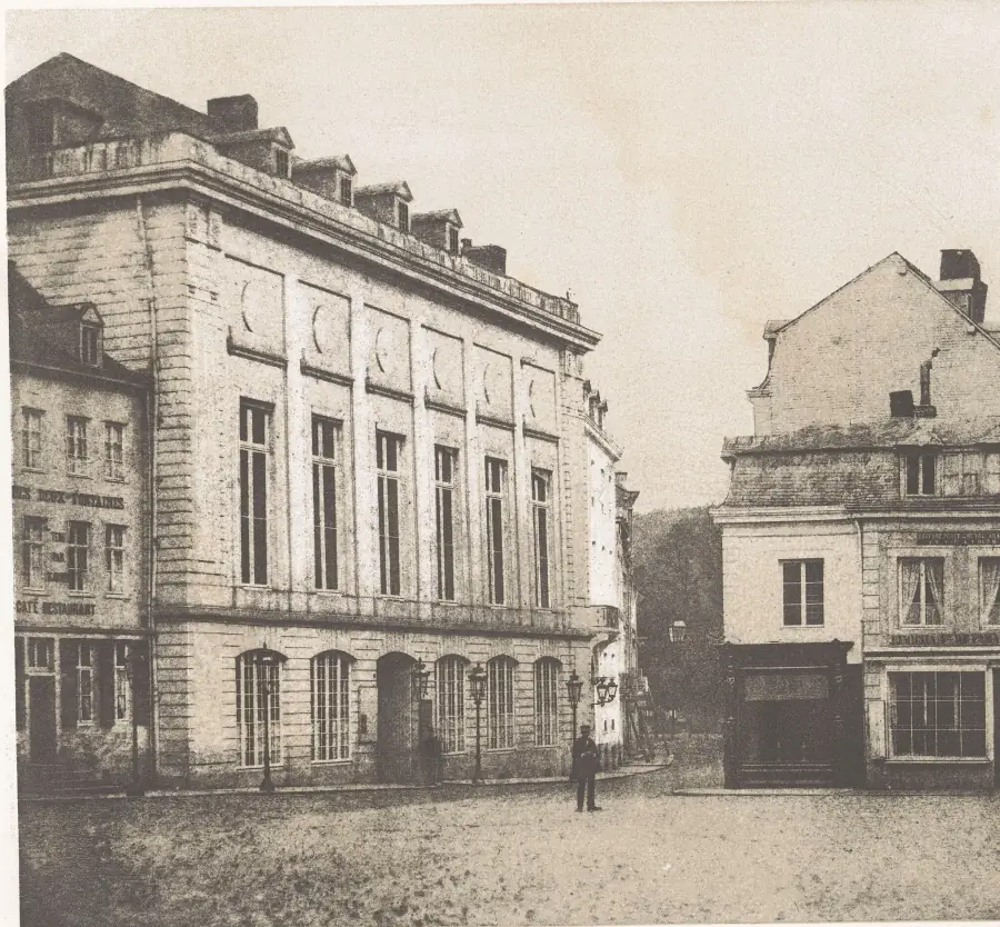 Photolithograph of La Redoute at Spa around 1860: a plain neoclassical block with tall arched ground-floor windows and oval medallions along the upper storey, a lone man standing on the empty square in front, a cafe and a gabled house to the right.