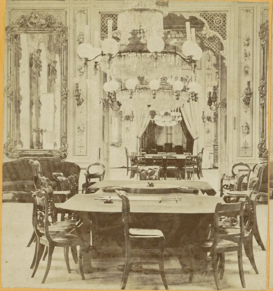 Half of a sepia stereoscopic card showing the gaming rooms of the old Wiesbaden Kurhaus: a long oval gaming table with a rack down its centre, bentwood chairs pushed back around it, gas globes and a crystal chandelier overhead, and a further table under a draped alcove beyond.